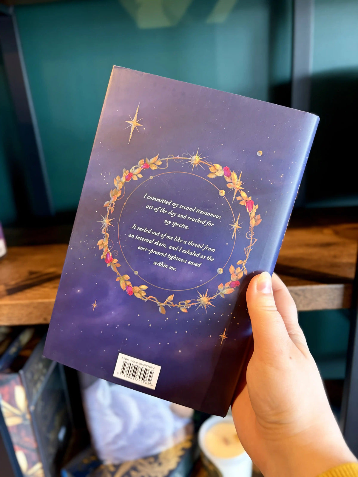 A hand holding the back of a paperback copy of the book 'Thorn Season' by Kiera Azar. The book cover features a dark purple background with a golden and red wreath, stars, and white text.