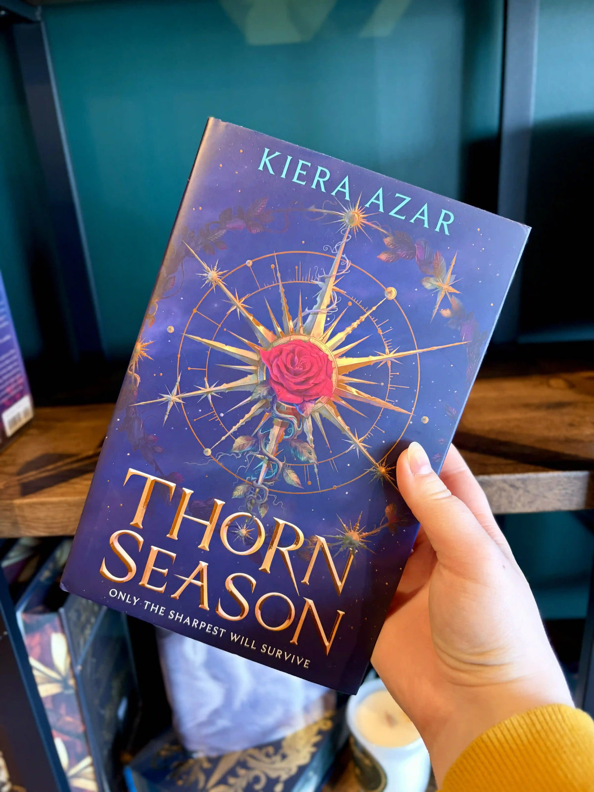 A hand holding a paperback copy of the book 'Thorn Season' by Kiera Azar. The book cover features a dark purple background with a golden compass design, a red rose in the center, and gold lettering.