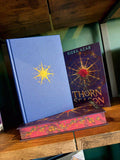 A hardcover book titled 'Thorn Season' by Kiera Azar displayed on a wooden shelf, standing upright next to another book with a blue cover and yellow starburst design.