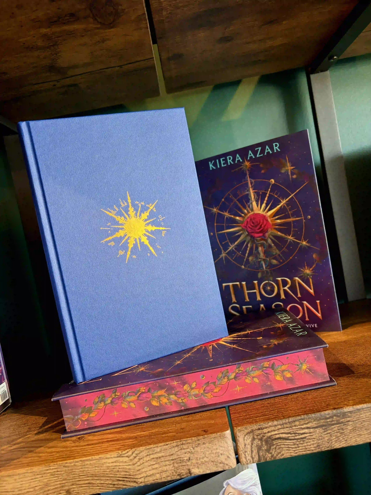 A hardcover book titled 'Thorn Season' by Kiera Azar displayed on a wooden shelf, standing upright next to another book with a blue cover and yellow starburst design.
