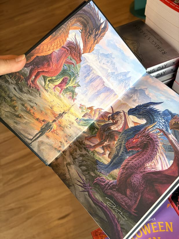 An open hardcover copy of 'Threshing Day' by Rebecca Yarros, showing the a fantastical landscape with two rows of dragons and humans walking between them.
