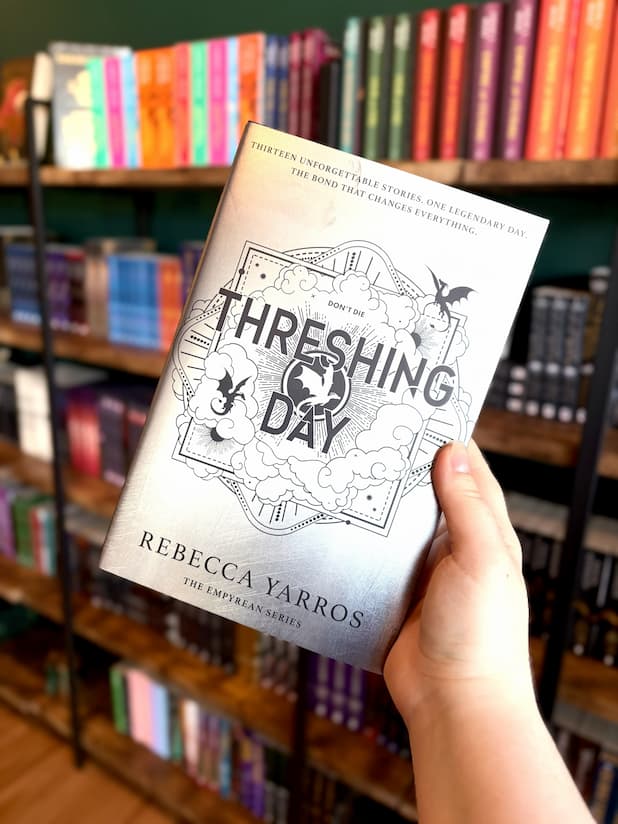 A hand holding a hardcover copy of 'Threshing Day' by Rebecca Yarros, showing the silver front cover and matching silver sprayed edges against a clean white background.