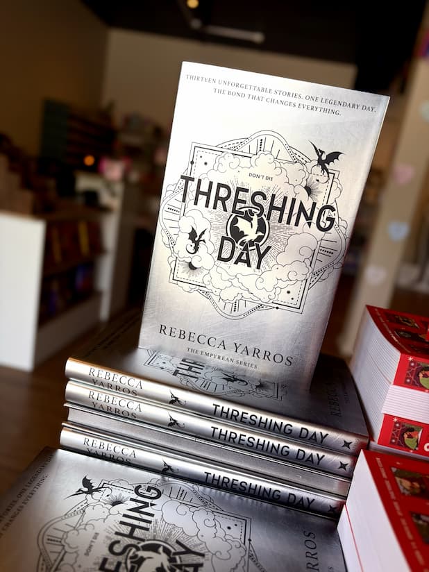 A stack of hardcover copies of 'Threshing Day' by Rebecca Yarros, showing the silver front cover and matching silver sprayed edges against a blurred bookshop background.