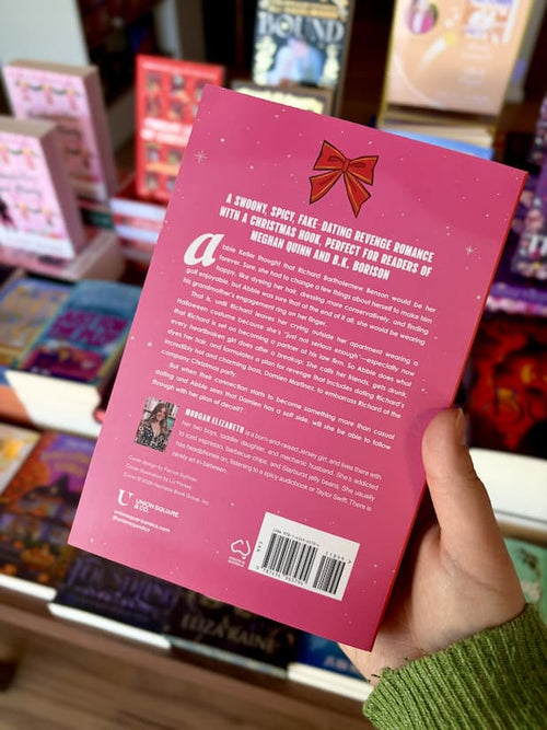 A hand holding the back of a copy of 'Tis the Season for Revenge' by Morgan Elizabeth, featuring a pink background, a red ribbon and white text.