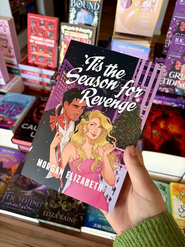 A hand holding a copy of 'Tis the Season for Revenge' by Morgan Elizabeth, featuring an illustrated couple in a snowy city scene at night with a Christmas tree and large white title text.