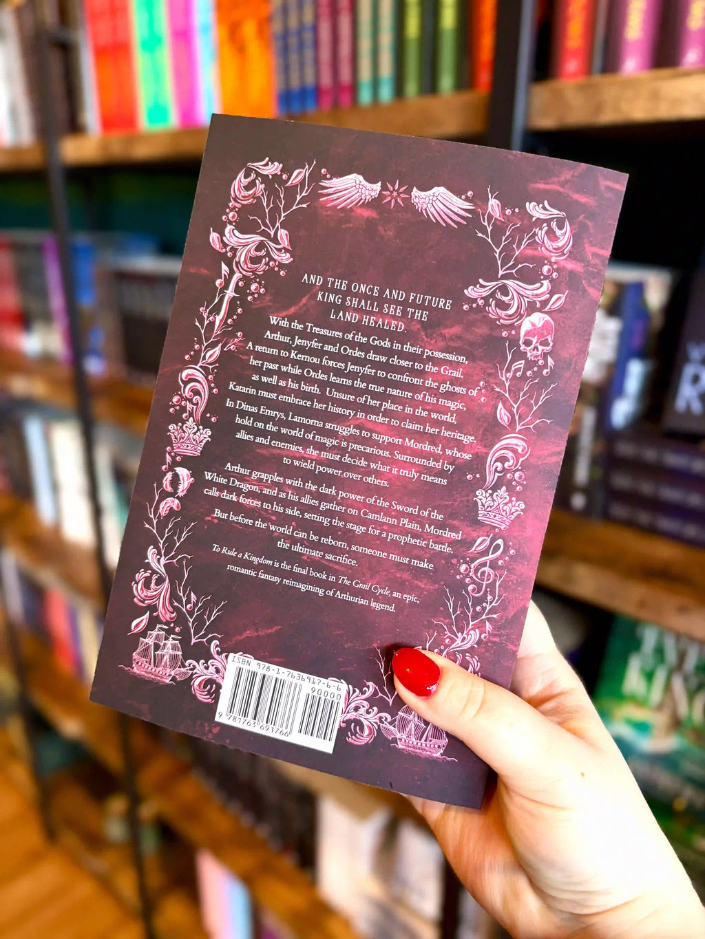 A hand with red nail polish holds the back of the book 'To Rule a Kingdom' by Kate Schmacher in a bookstore, with bookshelves blurred in the background. It is a crimson cover with a silver and white decorative border.