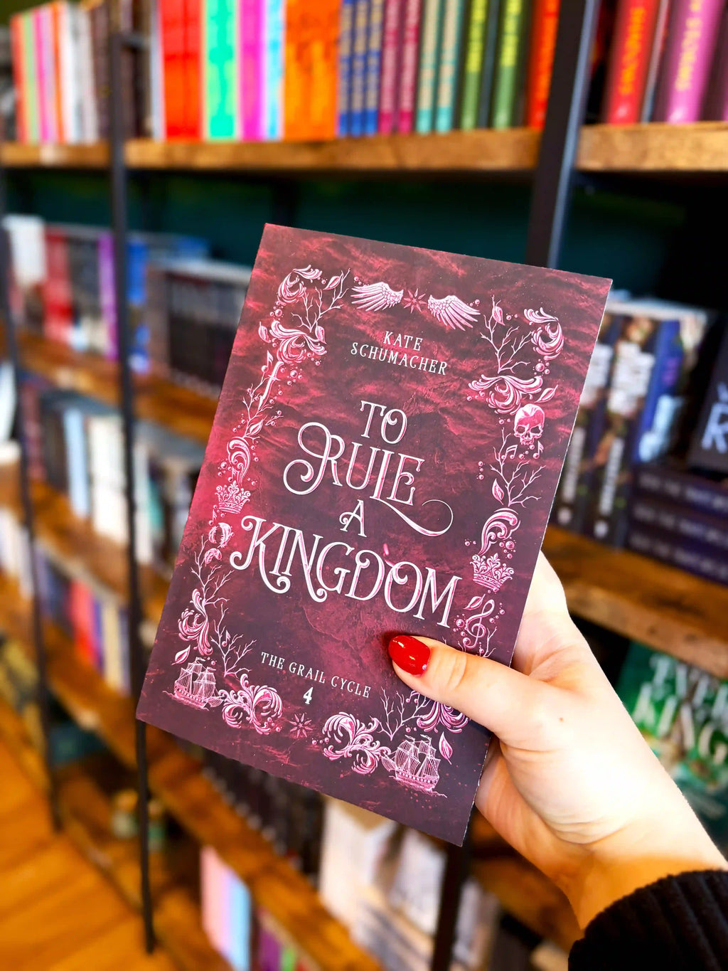 A hand with red nail polish holds a copy of the book 'To Rule a Kingdom' by Kate Schmacher in a bookstore, with bookshelves blurred in the background. It is a crimson cover with a silver and white decorative border and the title in the centre.