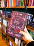 A hand with red nail polish holds a copy of the book 'To Rule a Kingdom' by Kate Schmacher in a bookstore, with bookshelves blurred in the background. It is a crimson cover with a silver and white decorative border and the title in the centre.