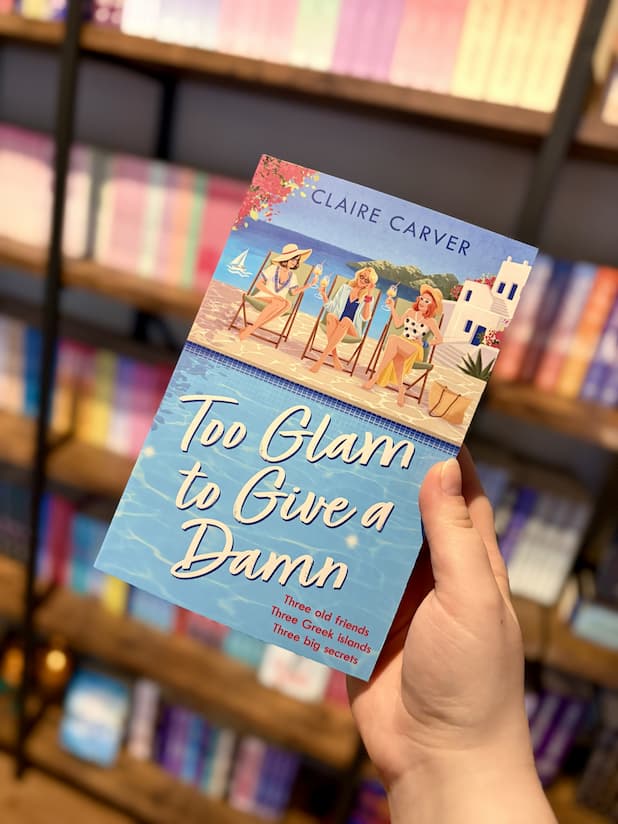 A hand holding a copy of 'Too Glam to Give a Damn' by Claire Carver with three women by a poolside drinking cocktails.
