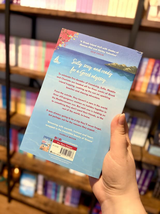 A hand holding the back of a copy of 'Too Glam to Give a Damn' by Claire Carver with a blue seaside background and white and red text.