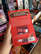 A hand with pink nail polish holding the back of a copy of 'The Twelve Loves of Christmas' featuring a bright red background with white text and windows with Christmas decorations.