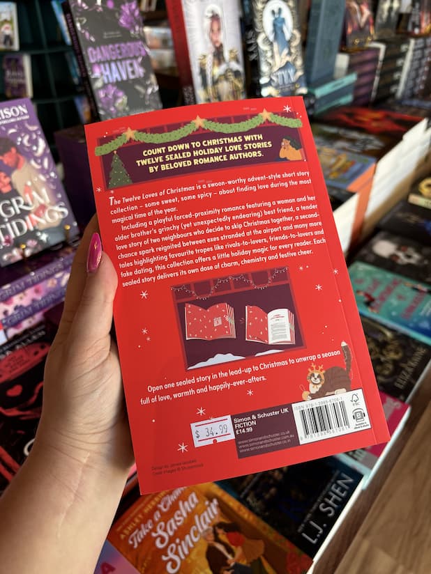 A hand with pink nail polish holding the back of a copy of 'The Twelve Loves of Christmas' featuring a bright red background with white text and windows with Christmas decorations.