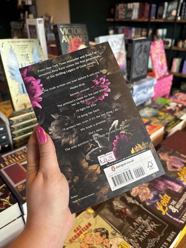 A hand with pink nail polish holds the back of the book 'Twisted Kingdom' by Rina Kent in a bookshop, with other book displays visible in the background. It is a dark cover with pink, grey, and yellow flowers with white text.