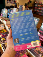 A hand holding the back cover of a book titled 'Two Left Feet' by Kallie Emblidge, with bookshelves visible in the background.