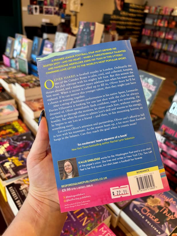 A hand holding the back cover of a book titled 'Two Left Feet' by Kallie Emblidge, with bookshelves visible in the background.