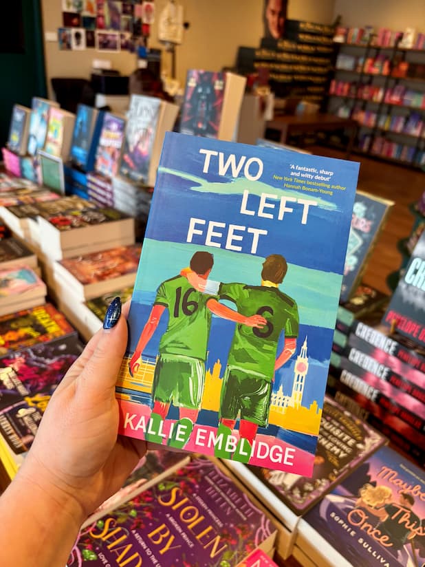 A hand holds the book 'Two Left Feet' by Kallie Embidge in a bookshop. The cover features an illustration of two soccer players in green jerseys standing on a beach with a city skyline in the background.