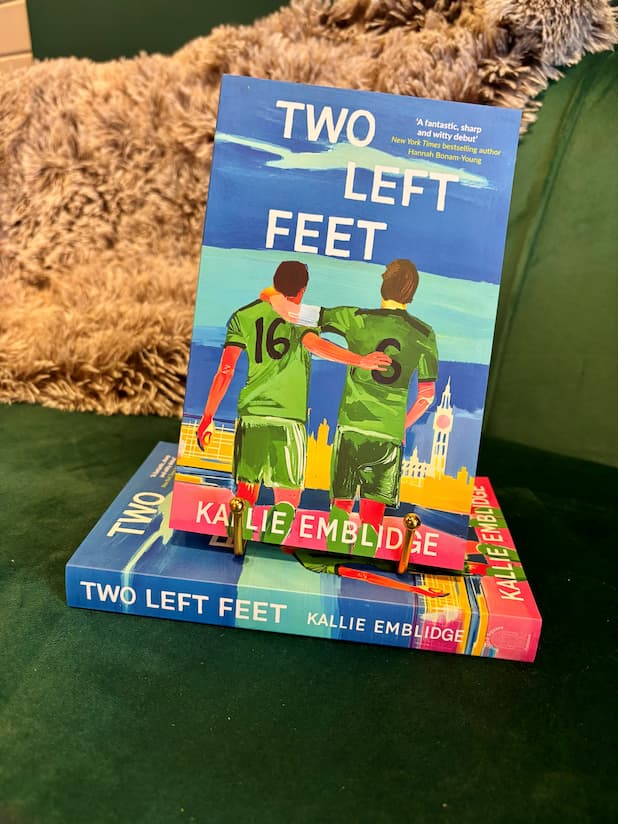 A book titled "Two Left Feet" by Kallie Emblidge is displayed upright on a teal couch, supported by two small gold bookends. The cover features an illustration of two soccer players in green jerseys standing on a beach with a city skyline in the background.