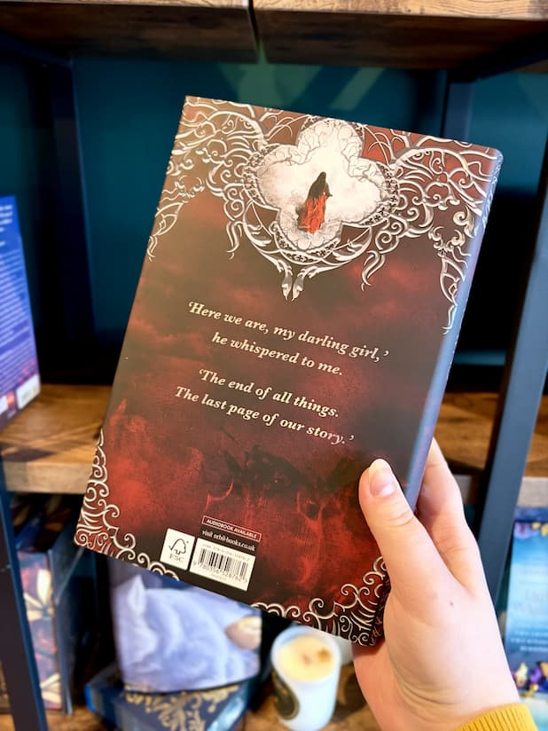 A hand holding the back of a hardcover book titled "Two Twisted Crowns" by Rachel Gillig. The cover features a red and brown background with ornate white filigree.