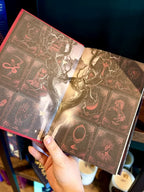 A hand holding a hardcover book titled "Two Twisted Crowns" by Rachel Gillig, showing the end pages. The pages are dark brown, black, red, and white with red symbols and a twisted tree.