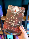 A hand holding a hardcover book titled "Two Twisted Crowns" by Rachel Gillig. The cover features a red and brown background with ornate white filigree and a small figure in red.