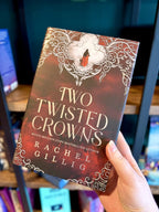 A hand holding a hardcover book titled "Two Twisted Crowns" by Rachel Gillig. The cover features a red and brown background with ornate white filigree and a small figure in red.