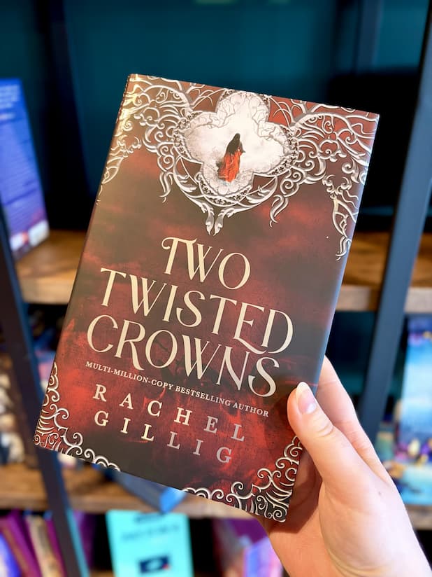 A hand holding a hardcover book titled "Two Twisted Crowns" by Rachel Gillig. The cover features a red and brown background with ornate white filigree and a small figure in red.