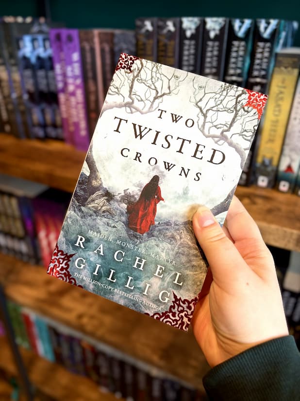 A hand holding a paperback copy of 'Two Twisted Crowns' by Rachel Gillig with a woman in a red coat walking through a foggy landscape.