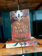 A hand holding a hardcover book titled "Two Twisted Crowns" by Rachel Gillig stands upright on top of a matching book on a wooden shelf. The cover features a red and brown background with ornate white filigree and a small figure in red.