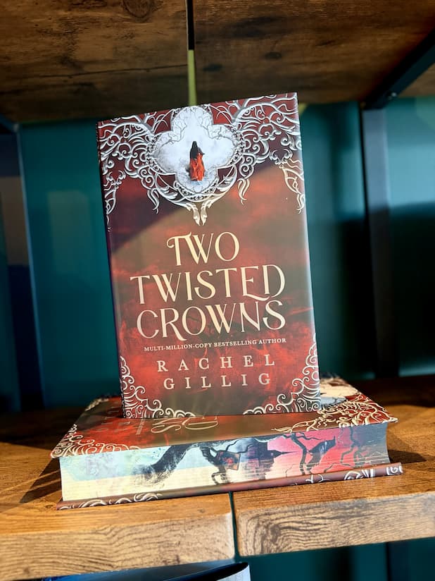 A hand holding a hardcover book titled "Two Twisted Crowns" by Rachel Gillig stands upright on top of a matching book on a wooden shelf. The cover features a red and brown background with ornate white filigree and a small figure in red.