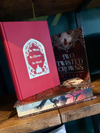 A hardcover book titled "Two Twisted Crowns" by Rachel Gillig stands upright on top of a matching book on a wooden shelf. The cover is red with silver text.