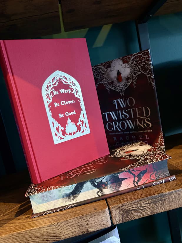 A hardcover book titled "Two Twisted Crowns" by Rachel Gillig stands upright on top of a matching book on a wooden shelf. The cover is red with silver text.