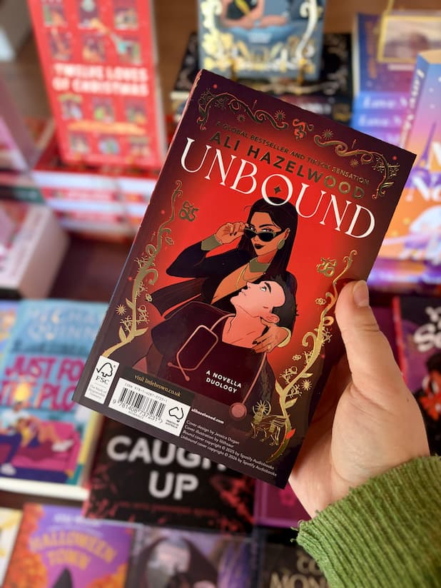 A hand holding a copy of the book 'Unbound' by Ali Hazelwood, featuring a red cover with gold border, a white title, and an illustrated couple embracing.