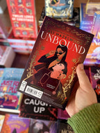 A hand holding a copy of the book 'Unbound' by Ali Hazelwood, featuring a red cover with gold border, a white title, and an illustrated couple embracing.