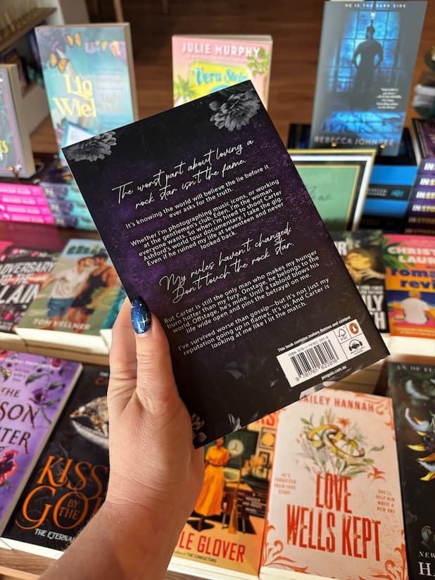 A hand holds the back cover of 'Unleashed by Eden' by Jade May from the Eden series, a spicy best friend's brother rockstar romance, with white font for the blurb on a purple background. The book is held in front of a table full of other blurry background. 