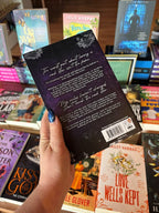 A hand holds the back cover of 'Unleashed by Eden' by Jade May from the Eden series, a spicy best friend's brother rockstar romance, with white font for the blurb on a purple background. The book is held in front of a table full of other blurry background. 