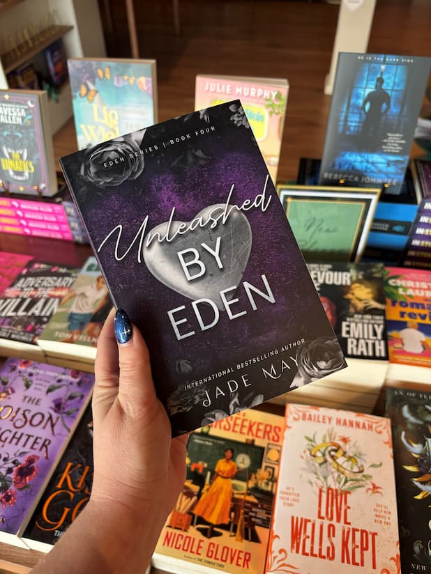 A hand holds the book cover of 'Unleashed by Eden' by Jade May from the Eden series, a spicy best friend's brother rockstar romance, with a silver heart and floral design on a purple background. The book is held in front of a table full of other blurry books in the background. 