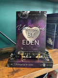 The book 'Unleashed by Eden' by Jade May from the Eden series sits on a gold stand on top of another book on a wooden bookshelf in front of a green wall. The book cover has a silver heart and floral design on a purple background.