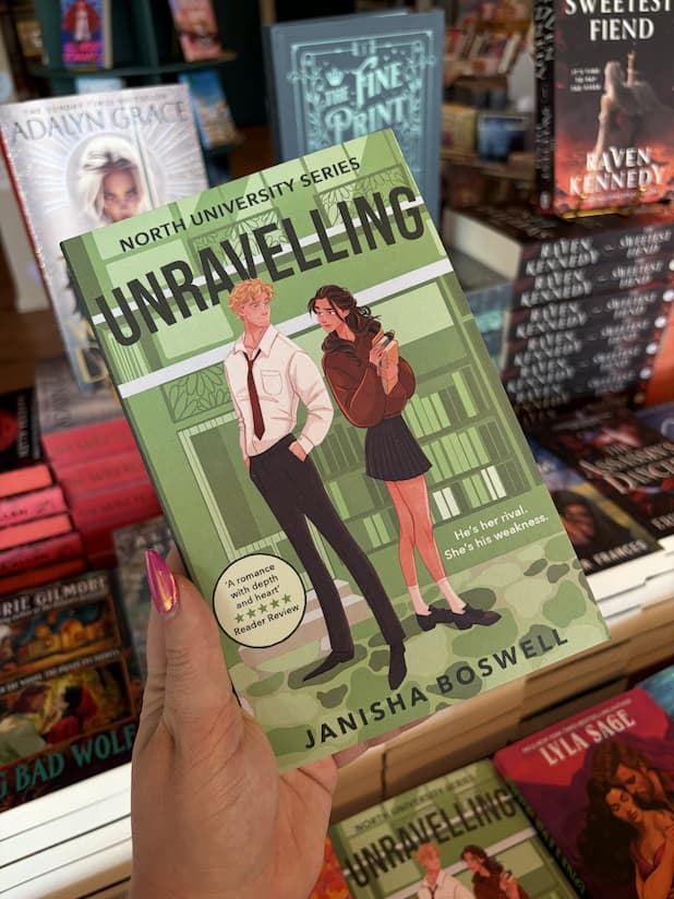 A hand with pink nail polish holds a copy of 'Unravelling' by Janisha Boswell, featuring two characters standing back-to-back and looking into each others eyes. They are standing against a green university background.