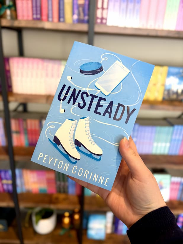 A hand holding a copy of 'Unsteady' with a graphic design of a hockey puck, ice skates, phone, earphones, and text on a blue background.