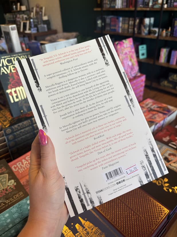 A hand with pink nail polish holding a paperback copy of 'Vengeful' by V.E. Schwab featuring vertical black lines drawn by a marker with black and red text.