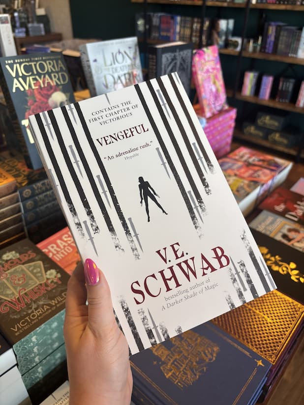 A hand with pink nail polish holding a paperback copy of 'Vengeful' by V.E. Schwab featuring vertical black lines drawn by a marker with grey daggers between the lines. In the centre of the page is the silhouette of a woman standing legs apart, holding two guns.
