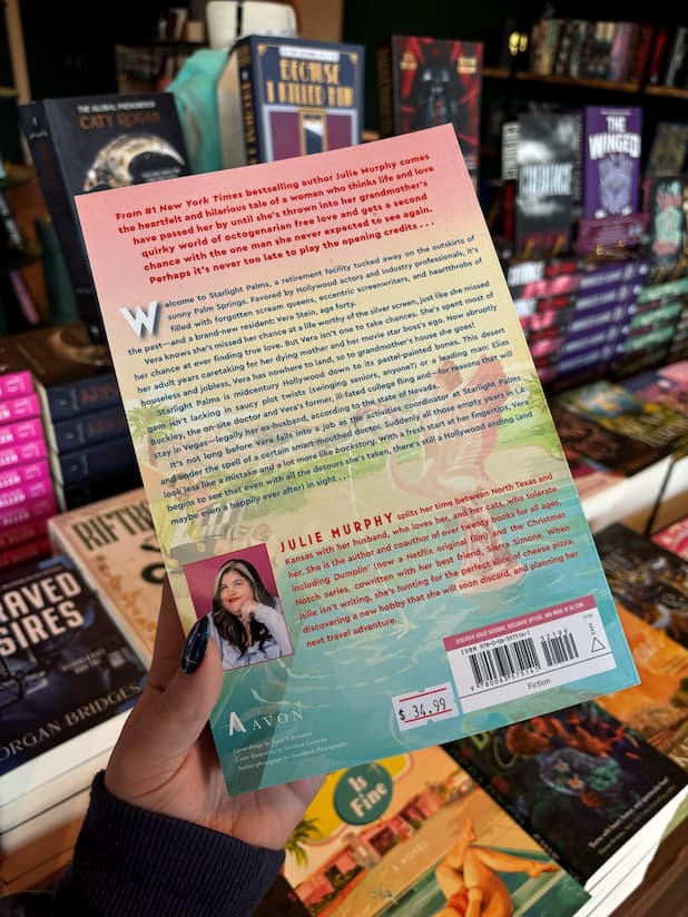 A hand holding the back cover of Vera Stein is Fine by Julie Murphy, with a background of other books on a wooden surface.