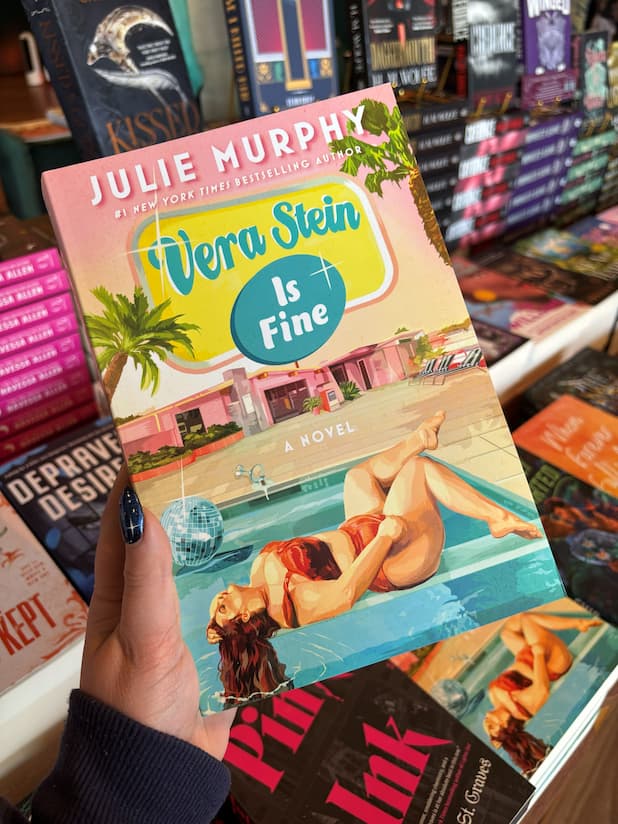 A hand with blue starry nail polish holds a paperback book titled 'Vera Stein is Fine' by Julie Murphy. The book cover features a quirky pink retirement village with a plus size woman in a bikini lounging by the pool.
