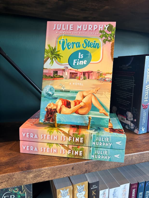 A copy of the book 'Vera Stein is Fine' by Julie Murphy displayed on a wooden bookshelf, standing upright on a small gold book stand next to other copies of the book.