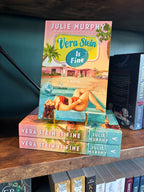 A copy of the book 'Vera Stein is Fine' by Julie Murphy displayed on a wooden bookshelf, standing upright on a small gold book stand next to other copies of the book.