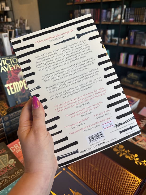 A hand with pink nail polish holding the back of a paperback cover of 'Vicious' by V.E. Schwab featuring lines of text overlaid with black marker and shapes of daggers and bone. In the middle is black and red text.