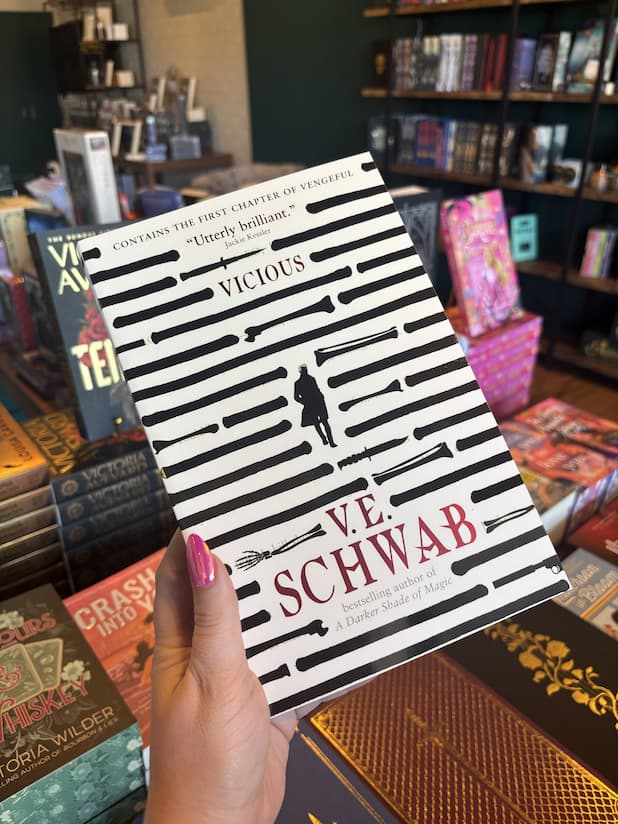 A hand with pink nail polish holding a paperback copy of 'Vicious' by V.E. Schwab featuring lines of text overlaid with black marker and shapes of daggers and bone. In the middle is the silhouette of a man n a trench coat.