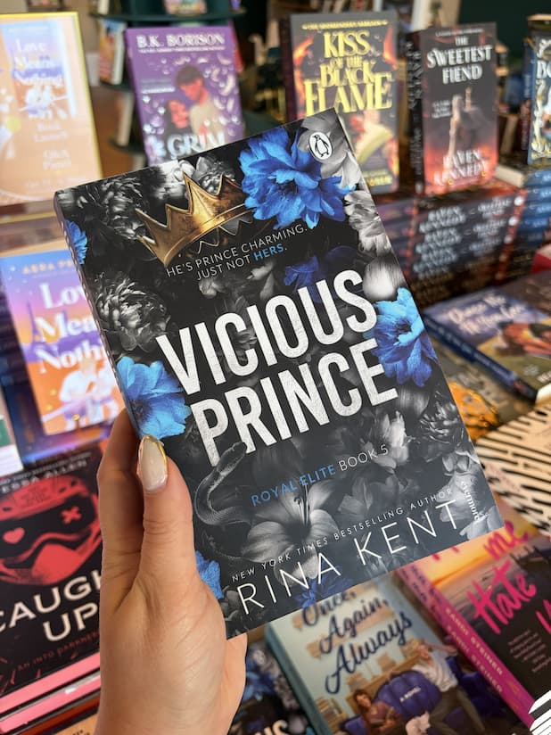 A hand with nude nail polish holds a copy of 'Vicious Prince' by Rina Kent, featuring a blue and silver floral design with large white text.