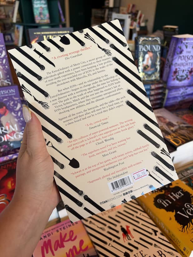 A hand with nude nail polish holding the back of a copy of 'Victorious' by V.E. Schwab featuring a pattern of bones, weapons and shovels, with black and red text.
