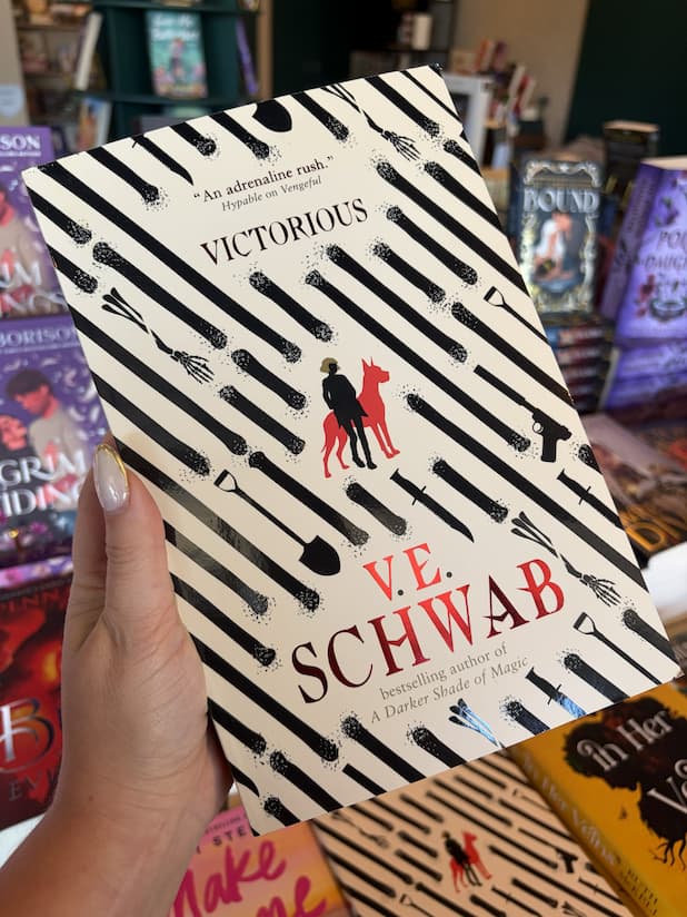 A hand with nude nail polish holding a copy of 'Victorious' by V.E. Schwab featuring a pattern of bones, weapons and shovels, with a silhouette of a person and dog at the centre.